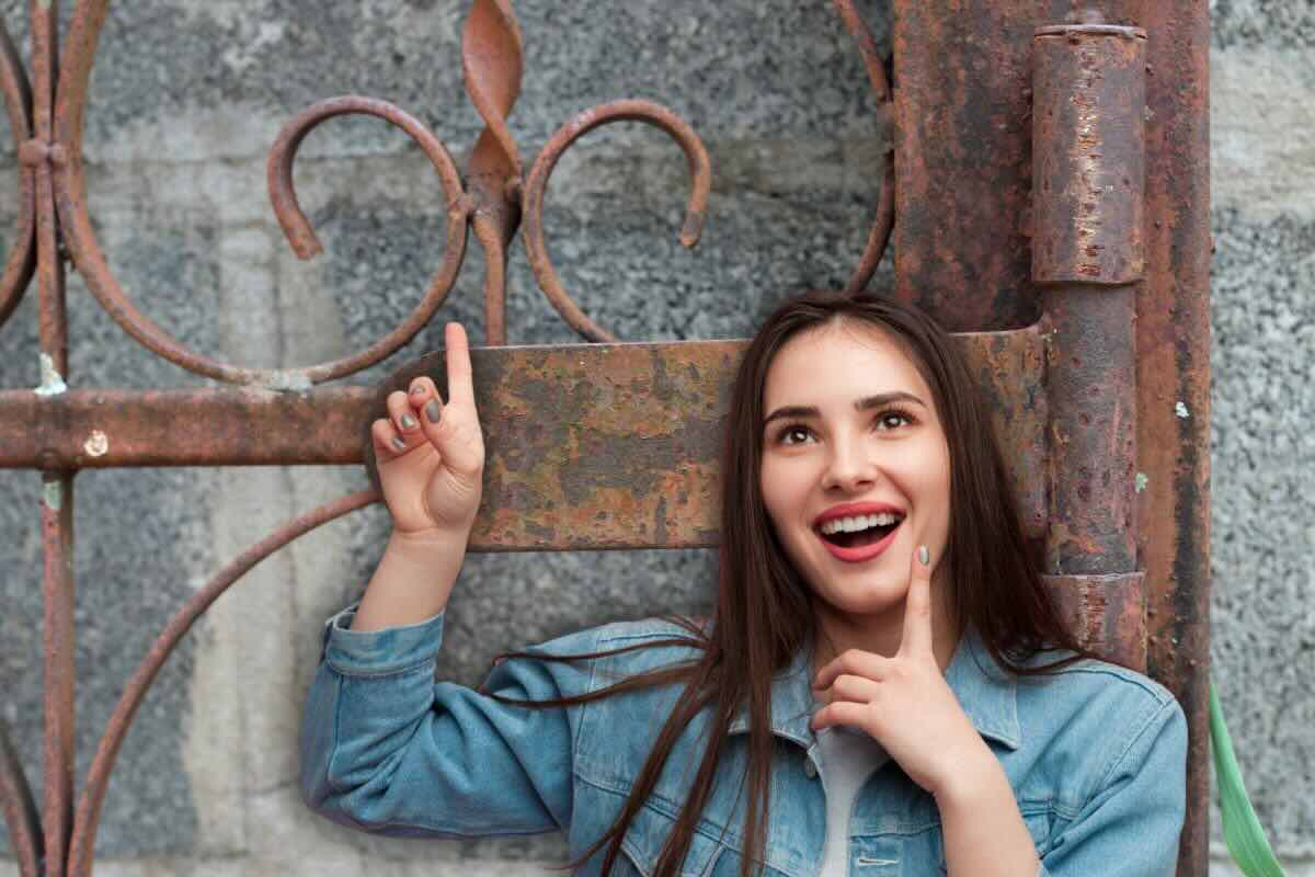Donna con dito alzato, sorridente, cancello dietro di lei pieno di ruggine