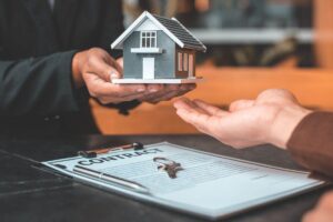 una persona che passa una piccola casa ad un'altra ed un foglio con scritto contratto