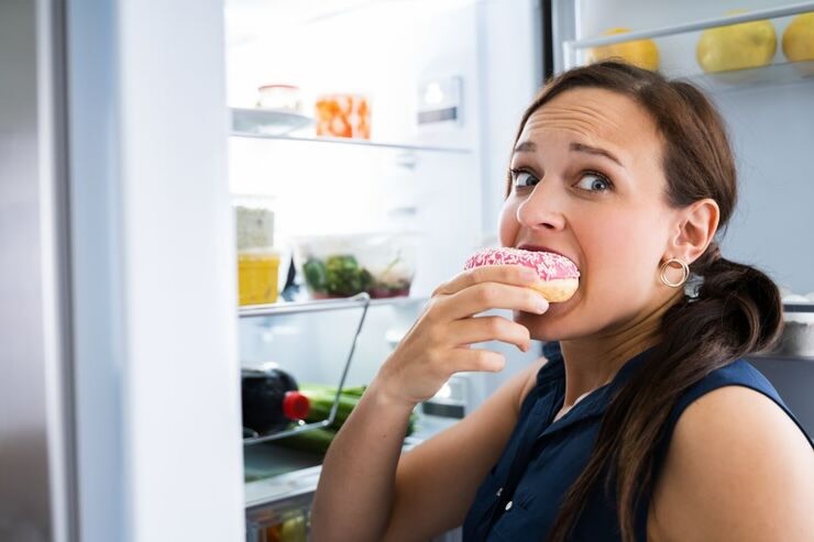 Donna mangia ciambella con glassa rosa vicino al frigo